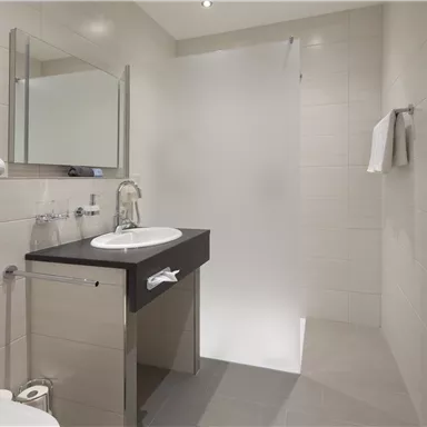 Ein modernes Badezimmer mit einer Dusche, einem Waschbecken und einem großen Spiegel. Die Wände sind hell und die Ausstattung ist schlicht und elegant.