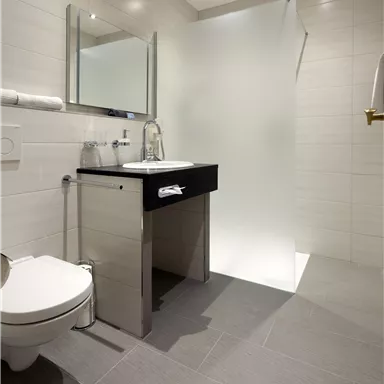 Ein modernes Badezimmer mit einem schwarzen Waschtisch, einem Spiegel und einer Duschkabine. Die Wände sind hell gefliest und der Boden ist grau.