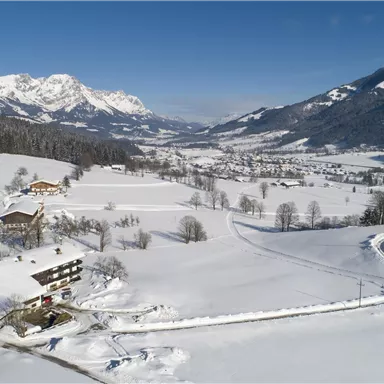 Eine verschneite Landschaft mit Bergen im Hintergrund. Bauernhäuser und ein ruhiger Weg verlaufen durch die Winterlandschaft.
