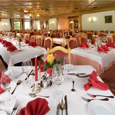 Ein eleganter Speisesaal mit vielen gedeckten Tischen. Rote Servietten und Blumenarrangements schaffen eine festliche Atmosphäre.