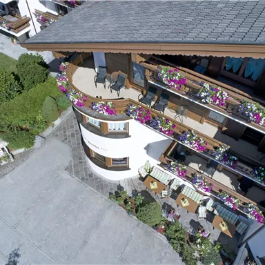 Ein charmantes Hotel mit schönen Balkonen, die mit bunten Blumen geschmückt sind. Im Vordergrund sind gemütliche Sitzgelegenheiten auf der Terrasse zu sehen.