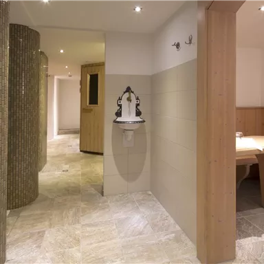 Ein eleganter Wellnessbereich mit modernen Fliesen und warmen Holzdetails. Auf der linken Seite befindet sich eine dekorative Säule, und ein Tisch mit Bänken lädt zum Verweilen ein.