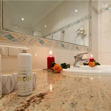 Ein Badezimmer mit einem weißen Waschbecken und einem großen Spiegel. Auf der Ablage stehen zwei Fläschchen sowie eine Kerze und eine Rose dekoriert.