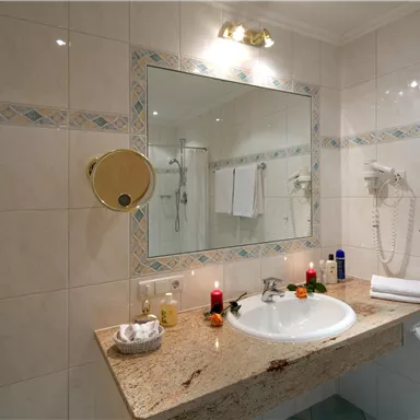 Ein modernes Badezimmer mit einem Waschbecken und einem großen Spiegel. Die Wände sind gefliest, und es gibt Handtücher sowie dekorative Kerzen.