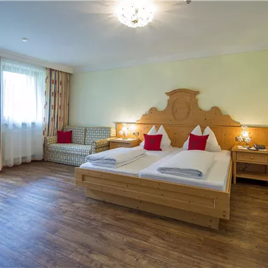 Ein gemütliches Hotelzimmer mit einem Doppelbett und zwei roten Kissen. Große Fenster sorgen für viel Tageslicht und eine angenehme Atmosphäre.