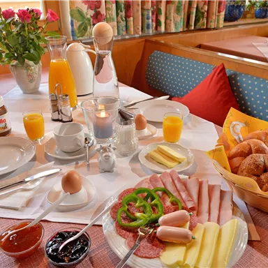 Ein reichhaltiges Frühstück mit Brötchen, Aufschnitt, Käse und Marmelade. Dazu gibt es frischen Orangensaft und Kaffee auf einem schön gedeckten Tisch.