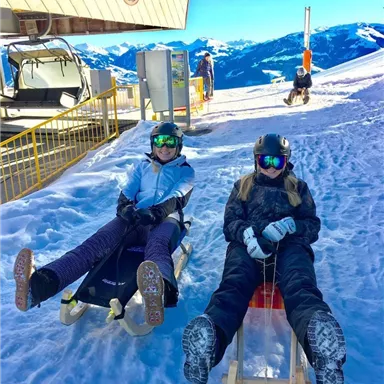 Zwei Personen sitzen auf Schlitten im Schnee, bereit für eine Abfahrt. Im Hintergrund sind Berge und ein blauer Himmel sichtbar.