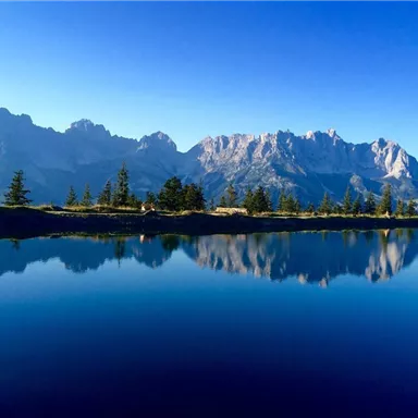 Eine malerische Berglandschaft mit klaren Wasserflächen und reflektierenden Gipfeln. Der Himmel ist strahlend blau und die Umgebung ist grün bewaldet.