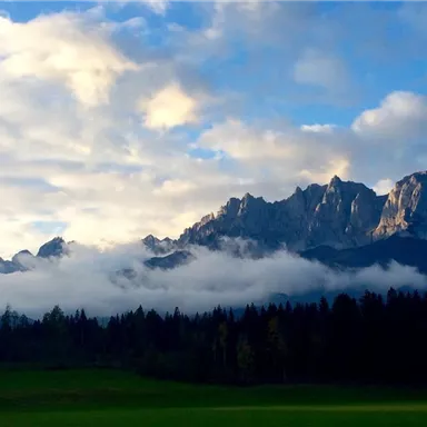 Eine beeindruckende Berglandschaft mit hohen Gipfeln und Wolken. Im Vordergrund erstreckt sich eine grüne Wiese.