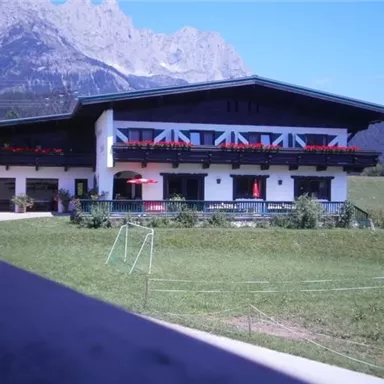 Ein traditionelles alpenländisches Gebäude mit Balkonen und roten Blumenbalkonen. Im Hintergrund sind Berge und ein grünes Feld zu sehen.