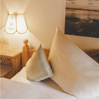 Ein gemütliches Schlafzimmer mit einem Bett, großen Kissen und einem Nachttisch. Eine Tischlampe sorgt für sanftes Licht und schafft eine entspannte Atmosphäre.