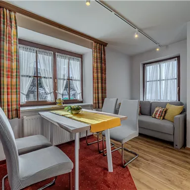 Ein gemütlicher Essbereich mit einem Tisch und vier Stühlen. Der Raum hat große Fenster mit karierten Vorhängen und einen hellen, einladenden Look.