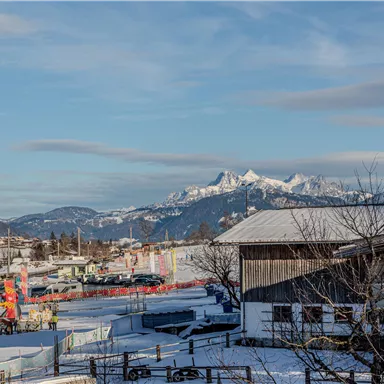 Eine verschneite Landschaft mit Bergen im Hintergrund und einem klaren Himmel. Im Vordergrund sind Gebäude und eine Skianlage zu sehen.