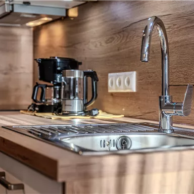 Eine moderne Küche mit einer Edelstahlspüle und einem Wasserhahn. Im Hintergrund steht eine Kaffeemaschine auf der Holzarbeitsfläche.