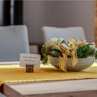 Ein schön gedeckter Tisch mit einer dekorativen Blumenschale auf einem gelben Tischläufer. Ein Willkommensschild steht ebenfalls auf dem Tisch.