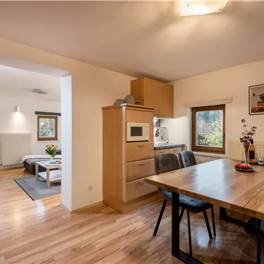 A modern kitchen with a large wooden table and stylish chairs. In the background are cozy living areas with windows that let in daylight.
