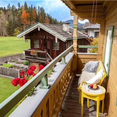 A balcony with a yellow table and a chair, offering a view of a green meadow and a traditional wooden house. Colorful flowers and trees in the background create a cozy atmosphere.