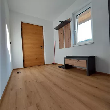 Ein heller Flur mit einem Holzfußboden und einer großen Tür. An der Wand sind Garderobenhaken und eine kleine Sitzbank.