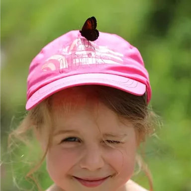 Ein lächelndes Mädchen mit einem Schmetterling auf ihrer pinken Kappe. Der Hintergrund ist grün und sonnig.