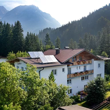 Ein modernes Haus im Gebirge, umgeben von grünen Bäumen und Bergen. Die Solarpanele auf dem Dach zeigen ein umweltfreundliches Design.