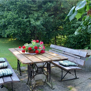 Ein schöner Garten mit einem Holztisch und Bänken. Auf dem Tisch steht ein Blumengesteck mit roten Geranien.