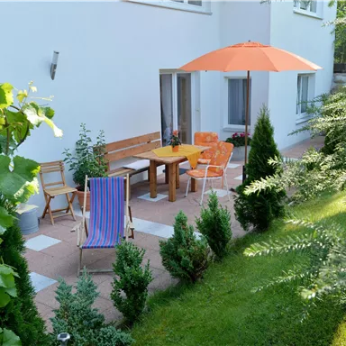 Eine gemütliche Terrasse mit einem Holztisch und Stühlen unter einem orangefarbenen Sonnenschirm. Umgeben von grünen Pflanzen und Rasen, ist der Platz ideal zum Entspannen.