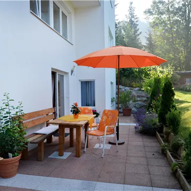 Ein gemütlicher Außenbereich mit einem Tisch, Stühlen und einem orangefarbenen Sonnenschirm. Umgeben von Pflanzen und einem gepflegten Garten.