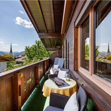 Ein sonniger Balkon mit gemütlichen Sitzgelegenheiten und einem Tisch. Im Hintergrund sind grüne Bäume und Berge zu sehen.