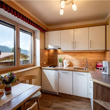 Eine helle Küche mit modernen Möbeln und viel Tageslicht. Auf dem Tisch stehen frische Früchte und eine Blumenvase.