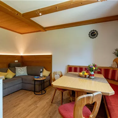 Ein gemütlicher Wohnraum mit einer grauen Couch und einem Essbereich. Am Tisch stehen frisches Obst und eine Flasche Wein.