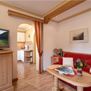 Ein gemütliches Wohnzimmer mit einem roten Sofa und einem Holztisch. Im Hintergrund sind eine Küche und ein Fernseher sichtbar.