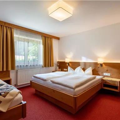 Ein gemütliches Schlafzimmer mit zwei Betten und warmen Holzakzenten. Das Zimmer hat ein großes Fenster mit Vorhängen und Teppichboden in Rot.