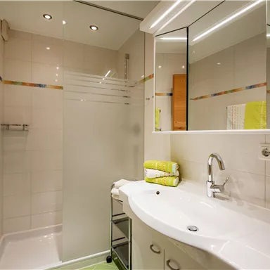 Ein modernes Badezimmer mit einer Dusche und einem Waschbecken. Die Wände sind hell gefliest, und es gibt farbige Handtücher und einen Spiegel.