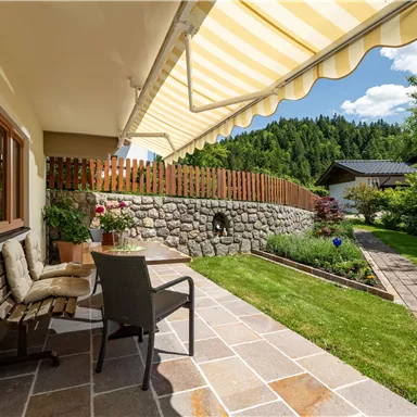 Ein einladender Außenbereich mit einem Tisch und Stühlen unter einem Sonnenschutz. Die grüne Gartenlandschaft ist von einer Steinmauer und Bäumen umgeben.