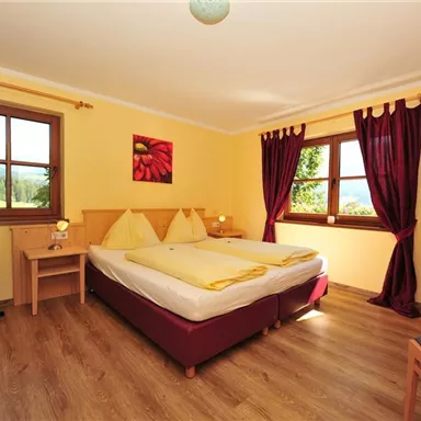 A cozy bedroom with a double bed and yellow walls. The windows offer a view of nature, and dark red curtains complement the decor.
