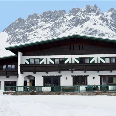 A large alpine-style house lies in the snow, surrounded by snow-covered mountains. It has an inviting porch and large windows.