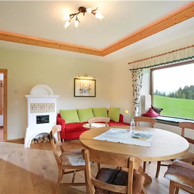 Ein helles Wohnzimmer mit einer gemütlichen Ecke und einem runden Holz-Tisch. Der große Fensterblick eröffnet eine grüne Landschaft.