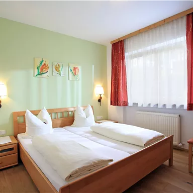 Ein gemütliches Schlafzimmer mit einem Doppelbett und einem hellgrünen Wandfarbe. Das Zimmer hat große Fenster mit Vorhängen und bietet eine einladende Atmosphäre.