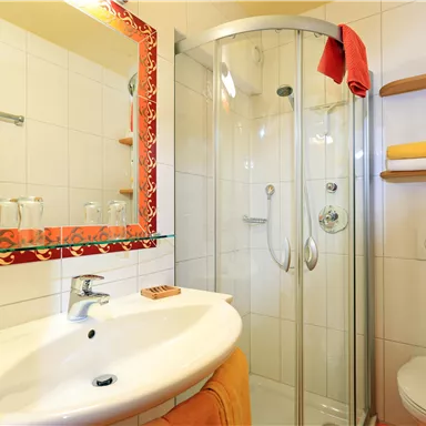 Ein modernes Badezimmer mit einer Dusche und einem Waschbecken. Die Wände sind hell gefliest und es gibt orangefarbene Handtücher.