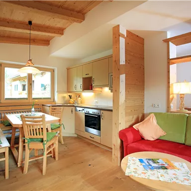 Eine helle, gemütliche Wohnung mit Holzbalkendecke. Die Einrichtung besteht aus einer Küchenzeile, einem Esstisch und einer Sitzecke mit einem roten Sofa.