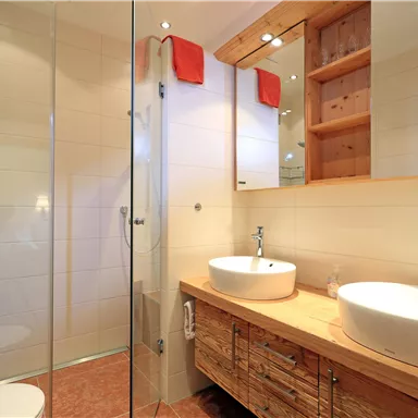 Ein modernes Badezimmer mit einer gläsernen Dusche und zwei Waschbecken. Die Möbel sind aus Holz und die Wände sind hell und freundlich gestaltet.