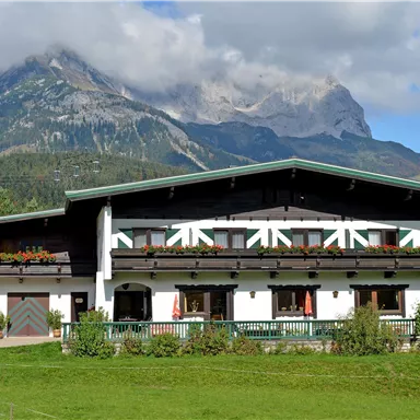 Ein traditionelles alpenländisches Haus mit schönem Blumenschmuck. Im Hintergrund erheben sich beeindruckende Berge unter einem klaren Himmel.