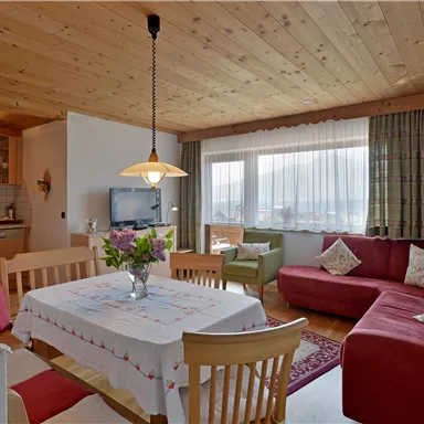 A cozy living area with a red sofa, a dining table, and large windows. The wooden beam ceiling and the flowers give the room a warm atmosphere.