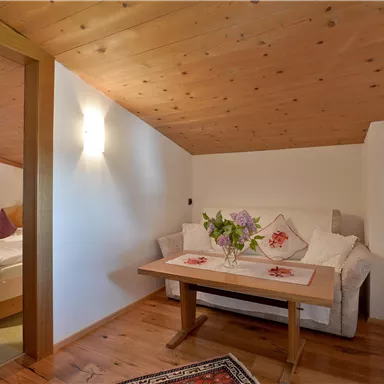 A cozy room with wooden ceilings and a table in the center. Fresh flowers are on the table and next to it is a sleeping area.