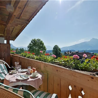 A cozy balcony with a view of the mountains. Breakfast platters are on the table and flowers are blooming in the boxes.
