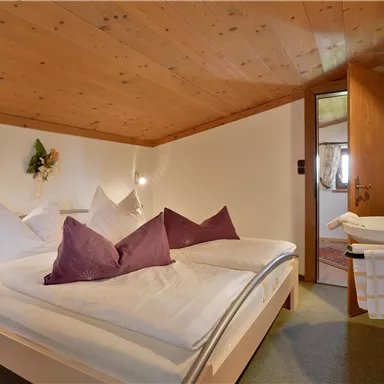 A cozy bedroom with a double bed and purple pillows. The wooden-paneled ceiling and soft lighting create a warm atmosphere.