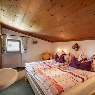 A cozy bedroom with a wooden attic. The room features a double bed with checkered blankets and an adjoining wash area.
