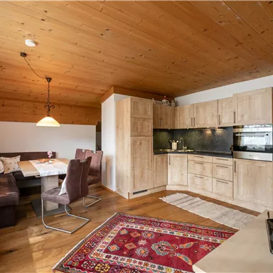 A cozy kitchen with wood details and a dining table for several people. The living space is bright and friendly with a rug and comfortable seating.