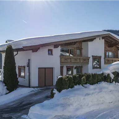 A beautiful house in alpine style, surrounded by snow-covered hills. The sun is shining under a clear sky.