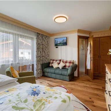 A bright room with two beds and a cozy sofa. Large windows offer views of the outdoors and wooden details give the space a warm atmosphere.